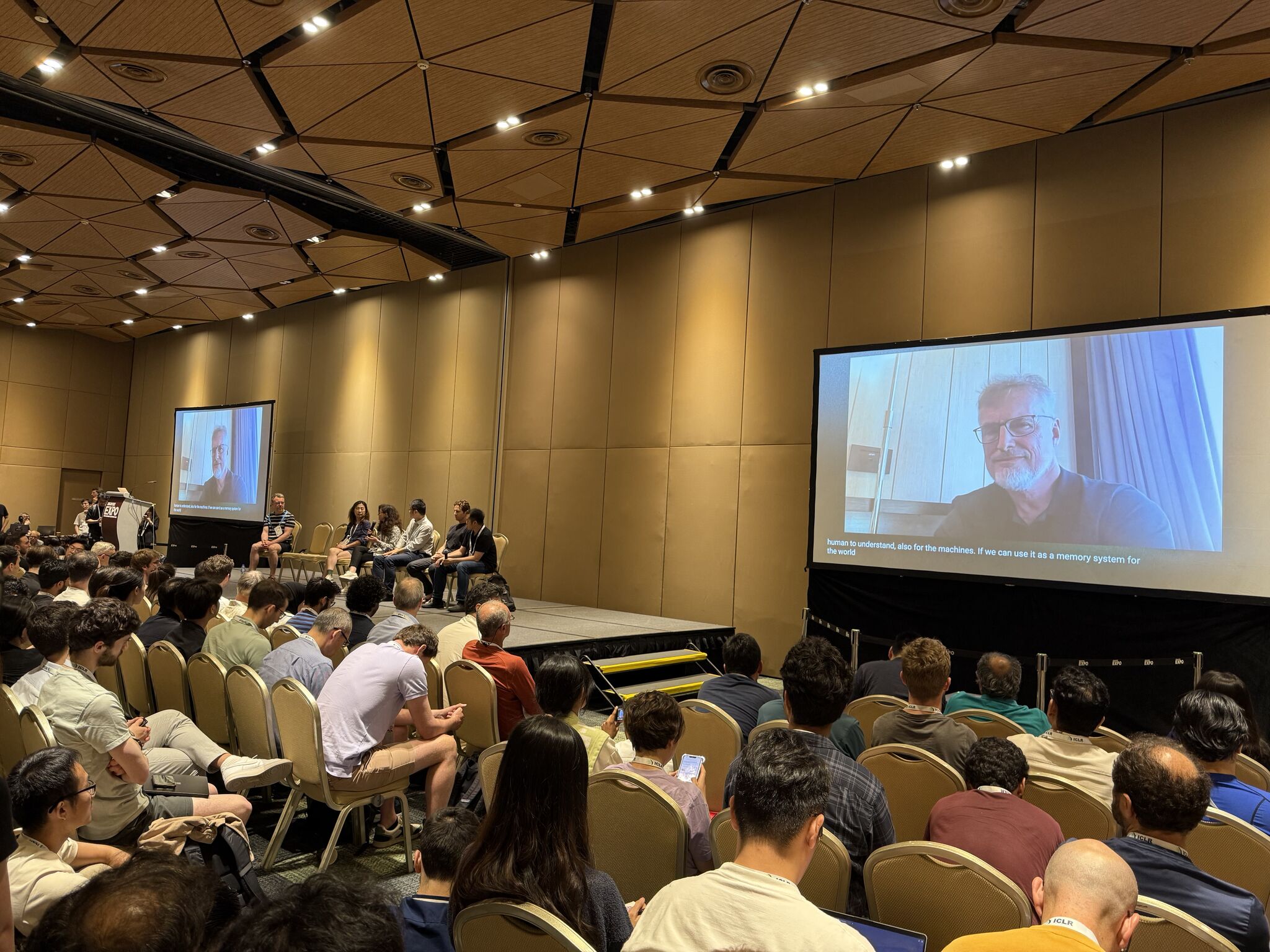 An audience attending a workshop panel discussion at ICLR 2025, with a remote speaker on screen.